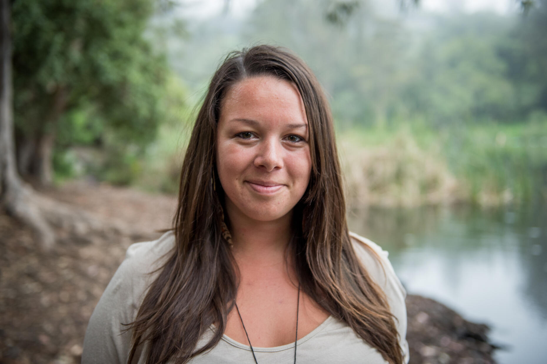 Headshot of Alisha Morton, a cis-gendered white woman with long brown hair, standing outdoors by a serene body of water with lush greenery in the background. She is wearing a light beige top, a necklace, and feather earrings, smiling gently in natural ligh